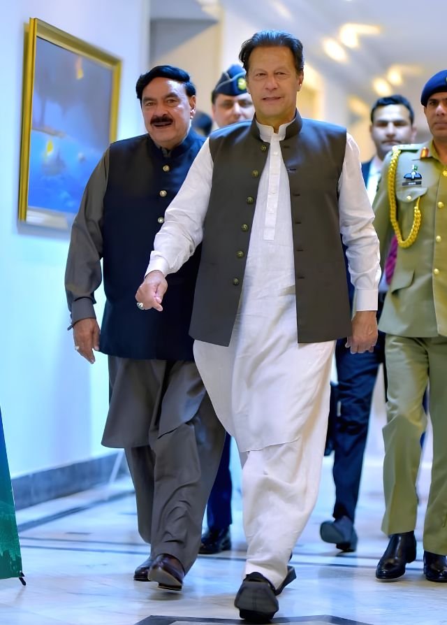 Former Prime Minister Imran Khan walking down a hallway alongside Sheikh Rasheed Ahmed and armed security officers in Islamabad.