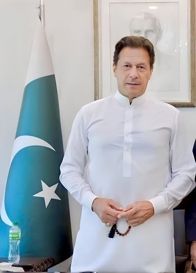 Former Prime Minister Imran Khan standing in a clean white shalwar kameez holding prayer beads with the Pakistan national flag and a framed portrait in the background.