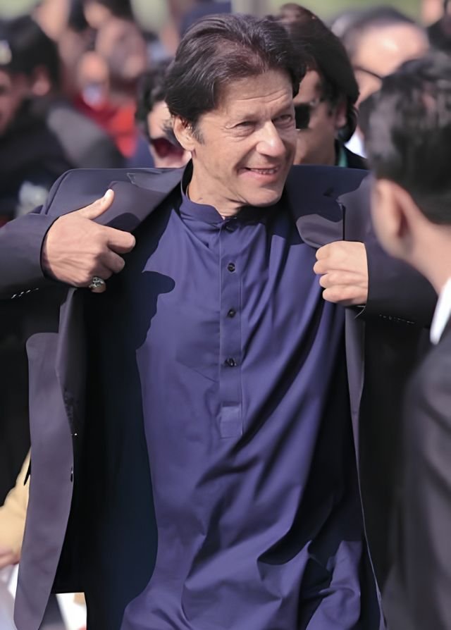 Imran Khan smiling and putting on his coat over a dark blue traditional shalwar kameez in a stylish outdoor setting.