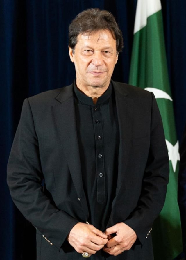 Formal portrait of Imran Khan wearing a traditional black sherwani with the Pakistani flag visible in the background.